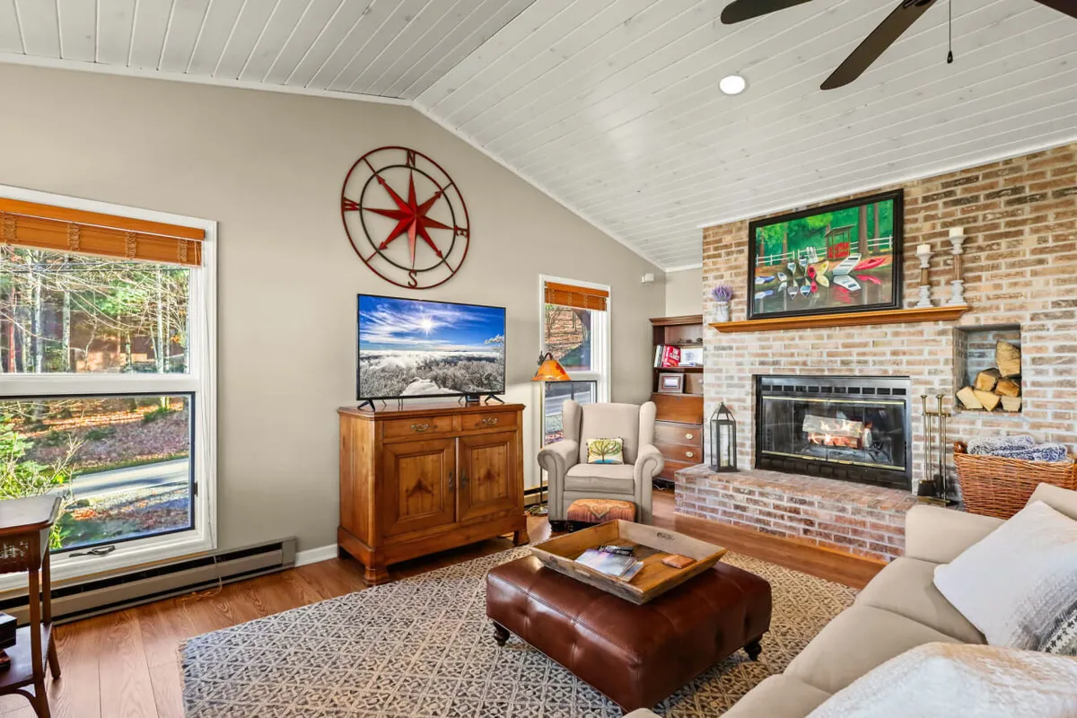 A warm and inviting living room featuring a fireplace, television, wooden cabinet, armchair, and decorative wall art. The room is filled with natural light during summer and offers a cozy atmosphere for winter evenings.