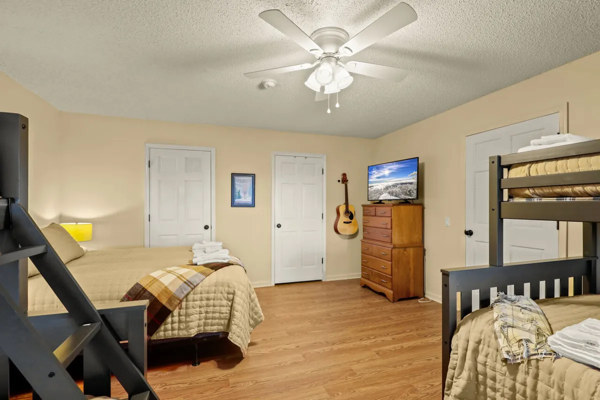 A spacious bunk room with a queen bed and a set of bunk beds. The room also includes a television, guitar, and dresser, suggesting both comfort and activities for families or groups. The décor and layout feel homey and relaxed, ideal for children or friends sharing the space.