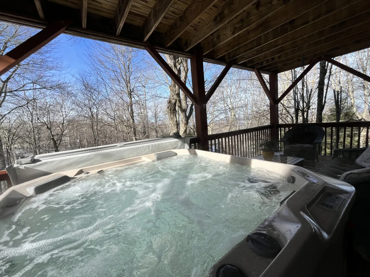 A spacious wooden deck with a hot tub as its centerpiece, surrounded by trees. The hot tub is positioned for relaxing while enjoying natural views, and there's additional seating for socializing or unwinding.