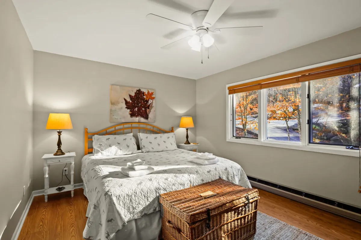 This image features a cozy bed with decorative linens, flanked by two bedside lamps, and a window letting in natural light. A woven storage basket sits nearby, lending to the tranquil, restful mood of the space. The overall ambiance is calm, inviting, and perfectly suited for relaxation.