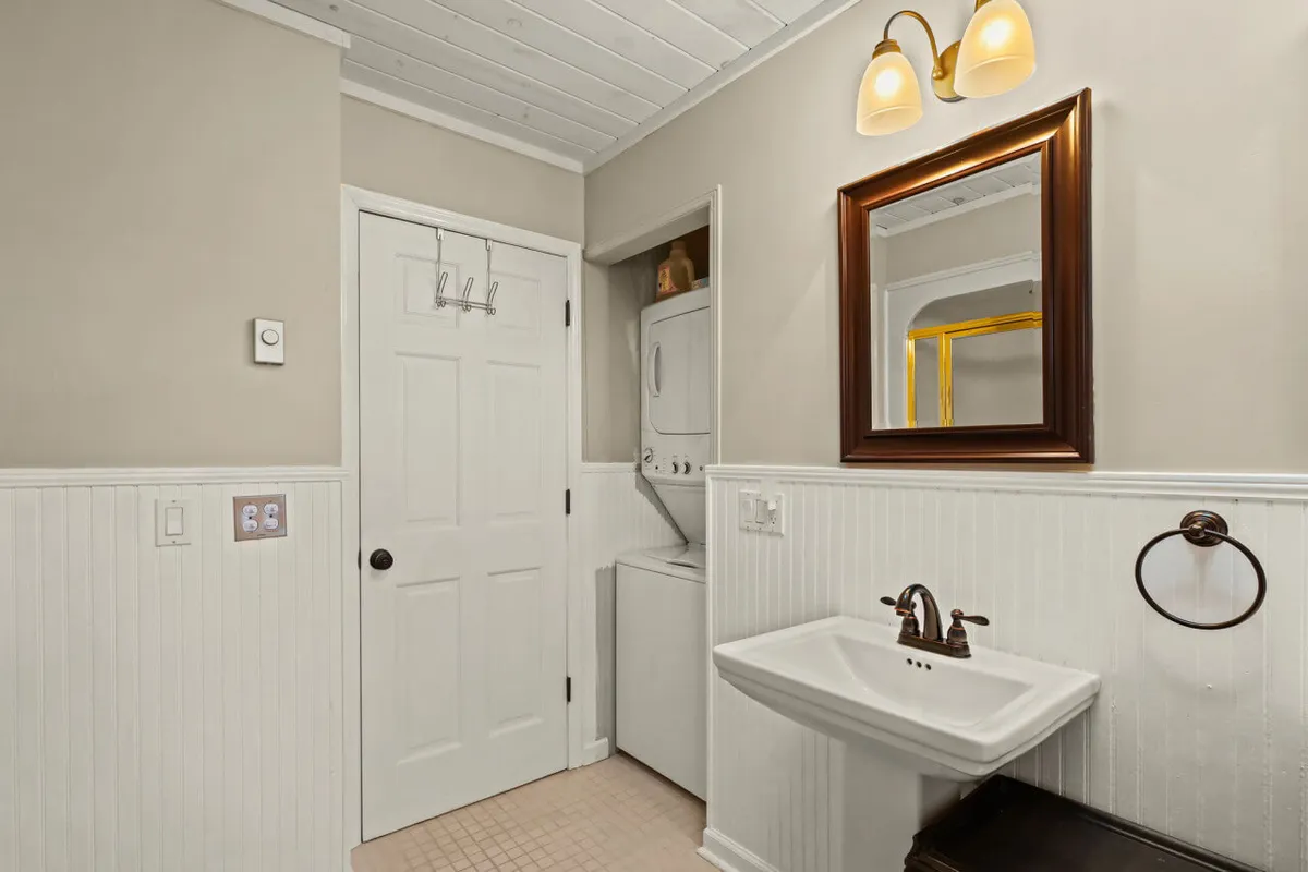 A practical and welcoming laundry area featuring a washer/dryer unit, a modern sink, and a mirror, all under bright lighting for easy use. The space feels fresh, organized, and conveniently located for guests to access.