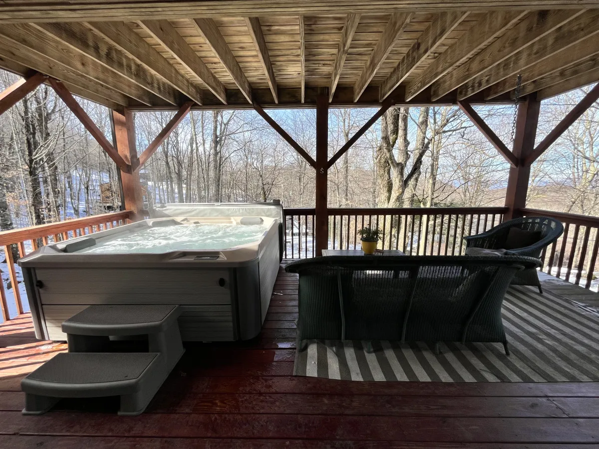 This image features a spacious outdoor hot tub set on a wooden deck, surrounded by lush trees. There is a comfortable seating area nearby, and the setting exudes tranquility with natural elements framing the space. The positioning of the hot tub offers sweeping sunset views and a sense of immersion in the mountain landscape.