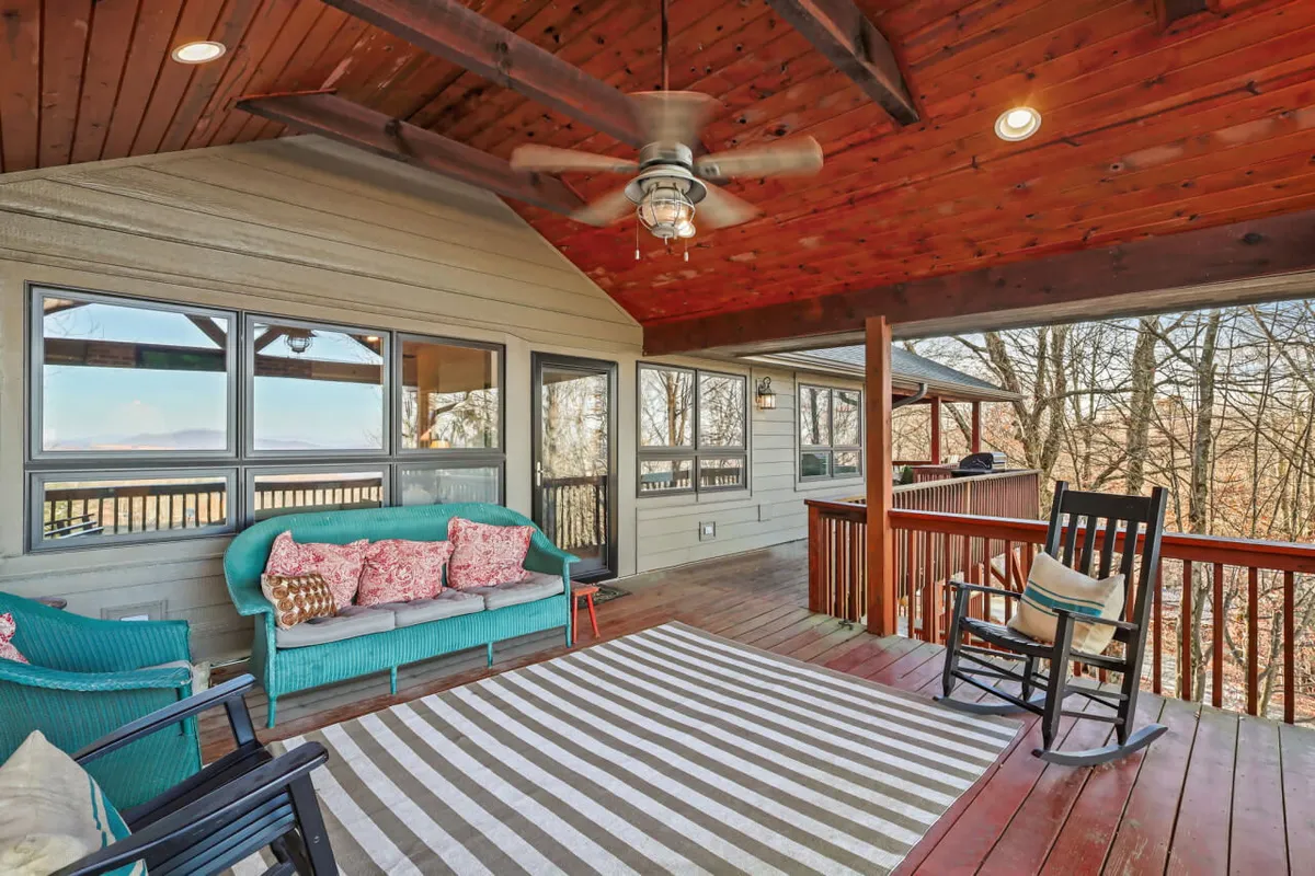 A cozy exterior seating area featuring a teal couch with cushions, a rocking chair, a striped area rug, and a wooden ceiling with exposed beams. The space is open to the view, inviting groups to gather, chat, and enjoy the fresh mountain air as the sun sets.