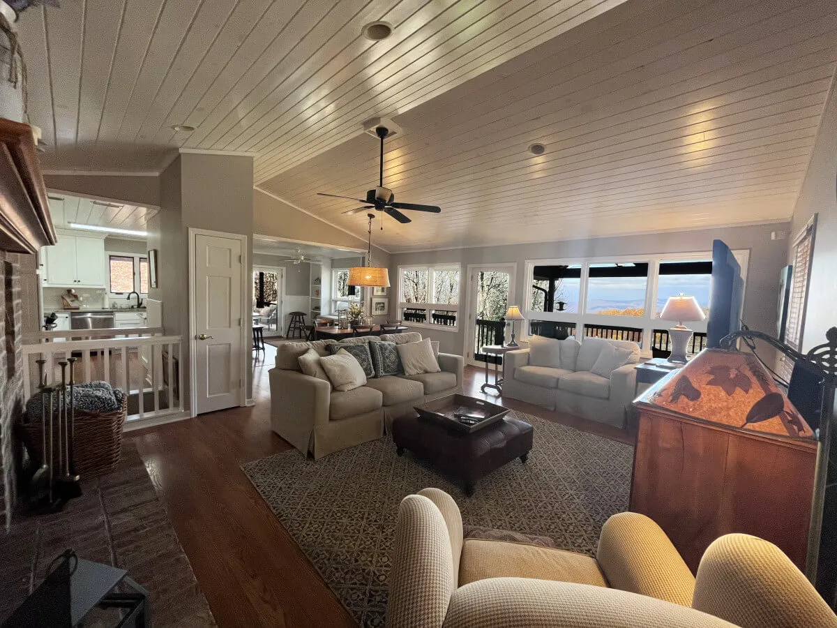 A welcoming interior view featuring a comfortable seating area with plush sofas, large windows that flood the space with natural light and showcase scenic views, a cozy fireplace, and an adjacent dining area—all arranged within an open-concept layout.