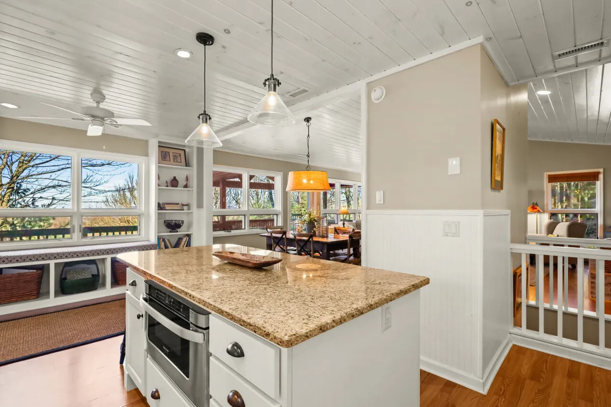 A bright, inviting kitchen and dining area with a granite-topped island, a spacious dining table, and large windows letting in cheerful daylight. The open flow between cooking and gathering spaces is evident, ideal for shared meals and conversations.