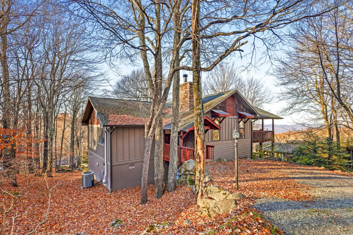 This exterior shot showcases The Spotted Fawn house nestled among mature trees with a prominent stone chimney. The home appears inviting and is surrounded by the vibrant colors of the changing seasons, reflecting the region's natural beauty.