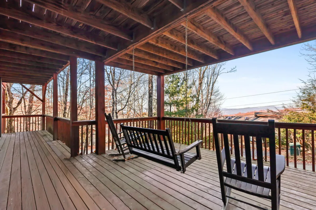 A wooden deck hosts a charming swinging bench, set against a backdrop of trees. The deck is elevated and open, providing peaceful sunset views and a connection to the changing foliage throughout the year. The mood is serene and contemplative.