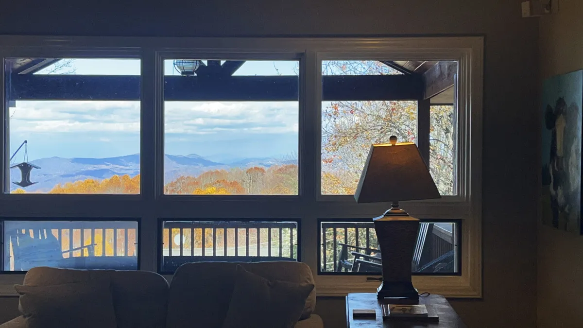 The hero image showcases a bright, inviting living area with large windows framing sweeping mountain views. A cozy sofa sits beside a glowing table lamp, offering the perfect perch to relax with a book or savor a morning coffee while sunlight pours in. The room exudes warmth and tranquility, inviting guests to unwind and soak in the beauty of Beech Mountain right from the comfort of their home away from home.
